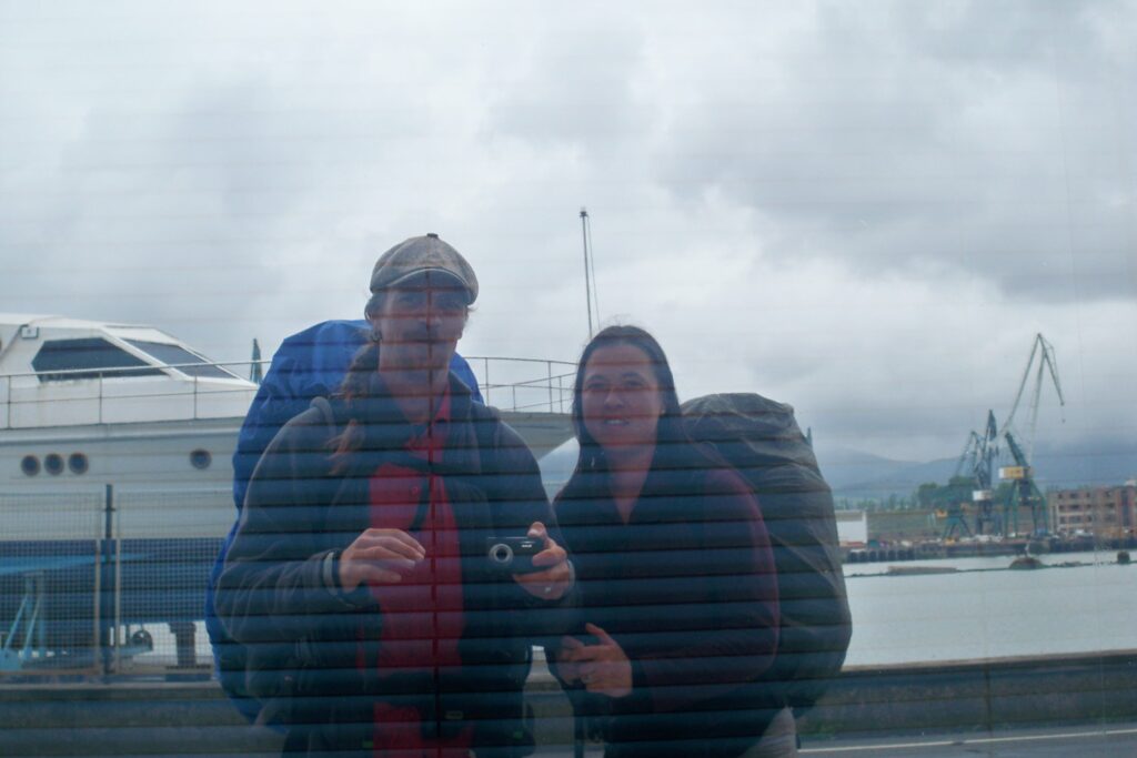 ein Mann und eine Frau spiegeln sich in einem Schaufenster am Kanal in Bilbao, beide tragen Wanderrucksäcke, Pilger, Selfie im Schaufenster, Pilgereise, Hafen