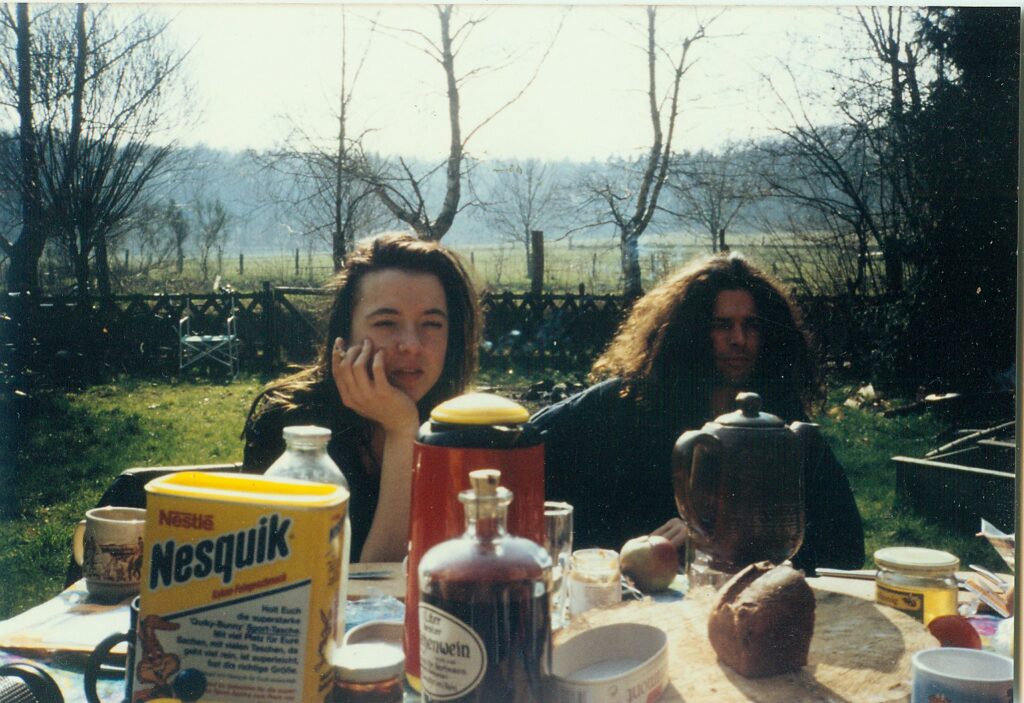 ein junge Frau und ein Mann mit langen Haaren sitzen im Garten am Frühstückstisch, es ist Herbst, auf dem Tisch stehen eine Kaffekanne, eine Teekanne mit Stöfchen, Brot, Honig, Kakao udn Käse