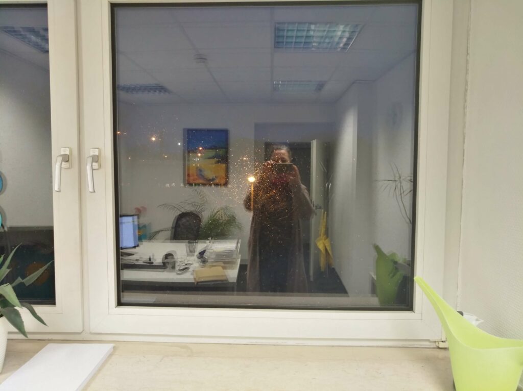 eine Frau fotografoert ihr eigenes Spiegelbild in einem Fenster, im Hintergrund ein Tisch mit einem PC und ein Bild an der Wand und eine geöffnete Tür