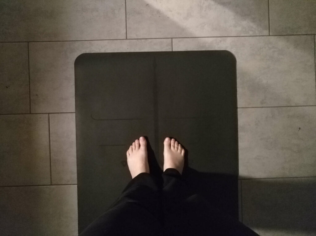 eine schwarze Yogamatte liegt auf einem Steinfußboden. Es sind Füße und untere Hosenbeine zu sehen