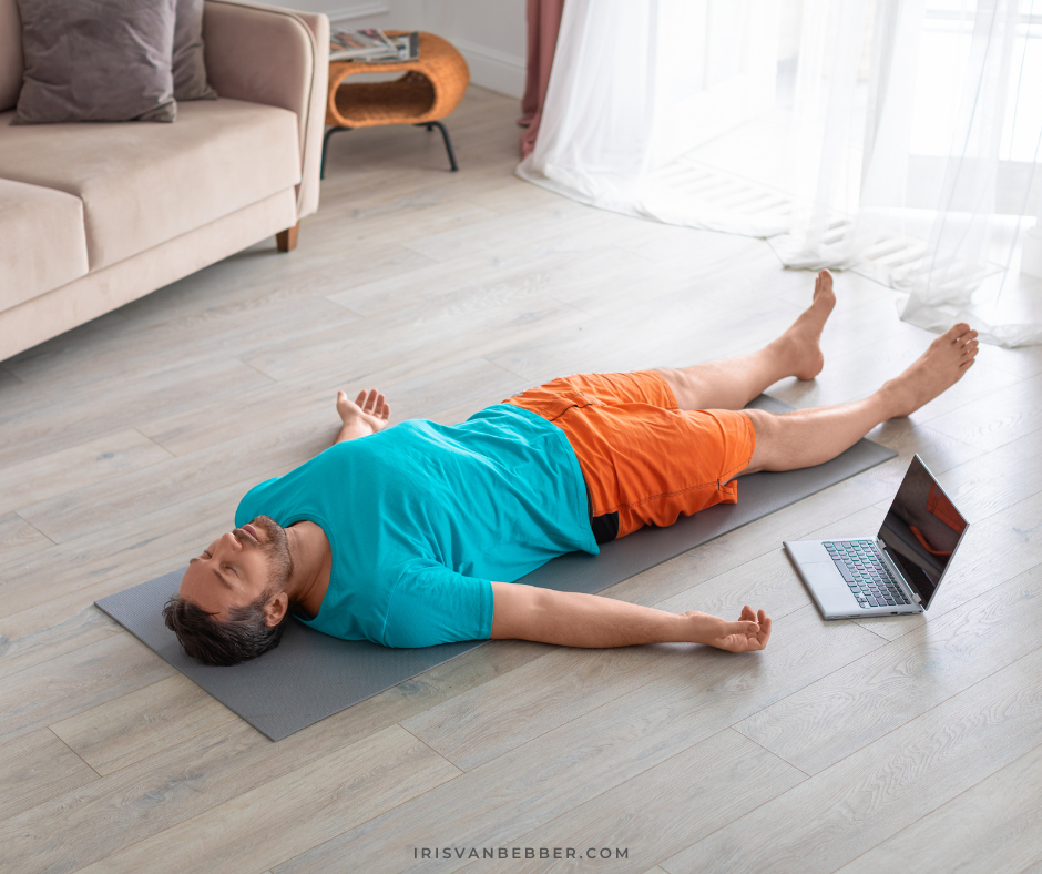 ein Mann liegt in einem Wohnzimmer auf einer Matte auf dem Rücken und übt Yoga Nidra, die Arme sind etwas zur Seite ausgestreckt, er hat die Aigen geschlossen, neben ihm steht ein aufgeklappter Laptop, er trägt eine kurze Hose und ein T-shirt