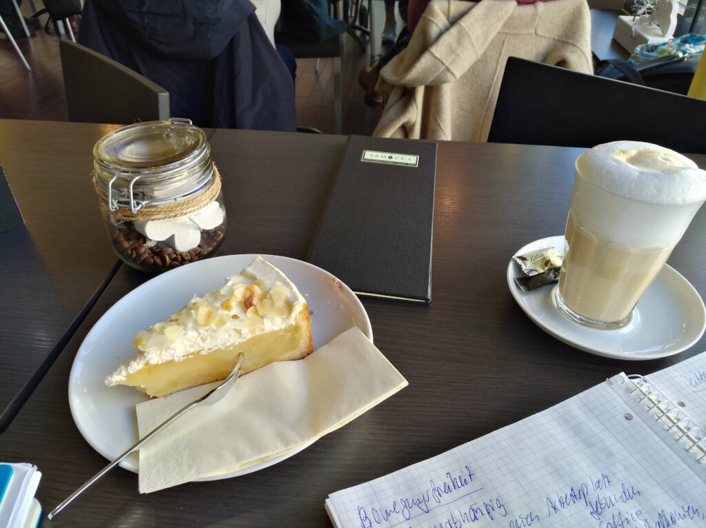 ein Tisch in einem Cafe mit einem Stück Apgelkuchen und einem Latte macchiato, daneben ein Blog mit handschriftlichen Ideen gesammelt für den Monatsrückblick Februar